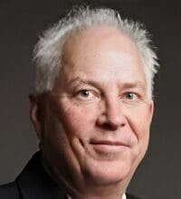 Portrait of an older man with short, gray hair wearing a dark suit, against a plain background.