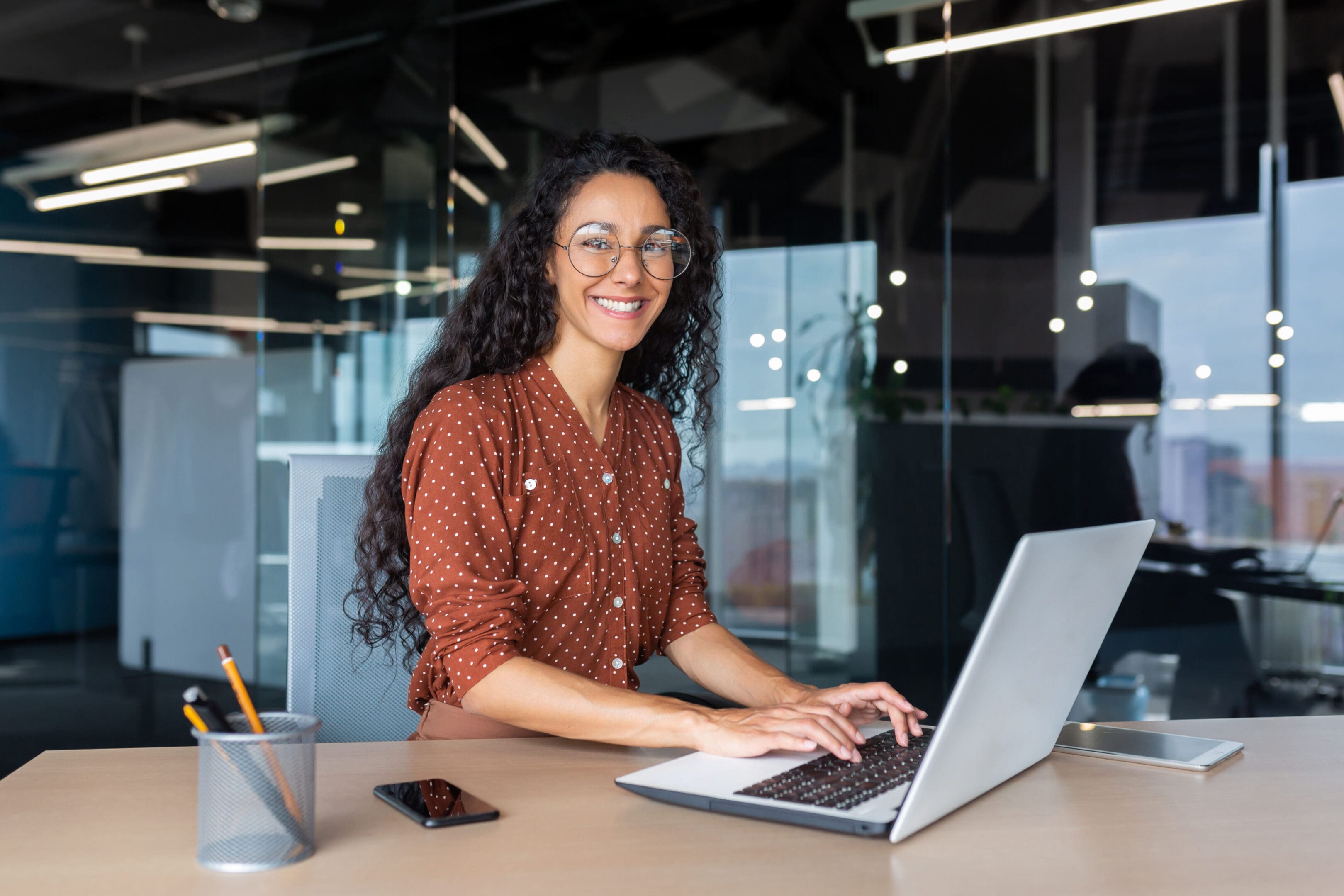 Person with long curly hair and glasses sits at a desk, smiling at a laptop. Office setting with a smartphone and pencil holder nearby.