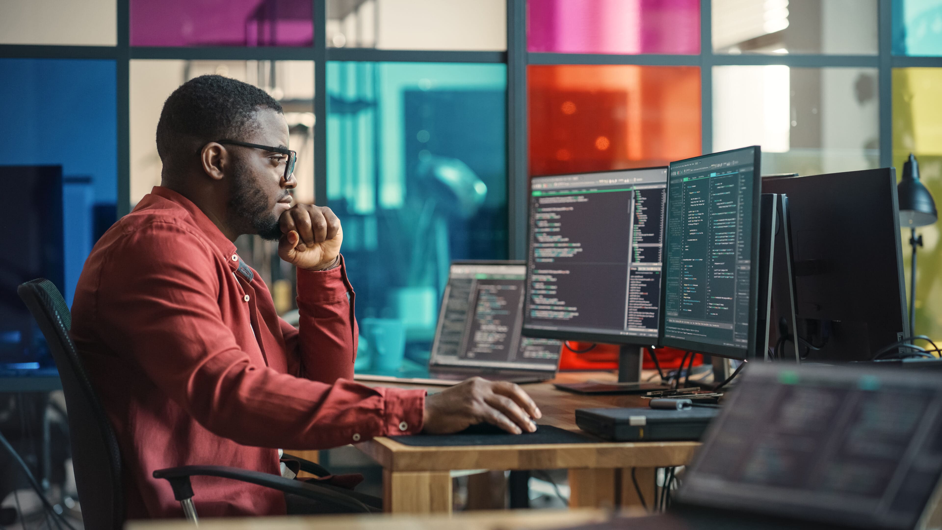 A person in a red shirt works intently on a desktop computer in an office with colorful window panels, surrounded by multiple monitors displaying code.