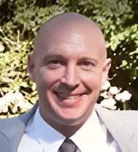 A person in a light suit and tie, smiling outdoors, with greenery in the background.