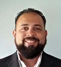 A bearded person smiling, wearing a dark suit jacket over a white collared shirt, against a plain light background.