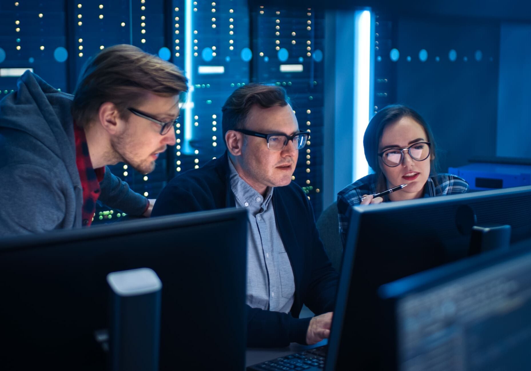 Three people in a dimly lit server room are focused on computer screens, working collaboratively.